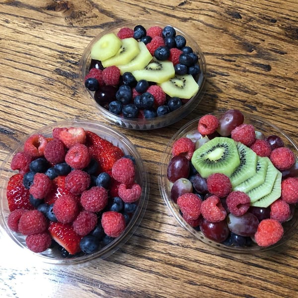 Three clear plastic containers filled with fresh fruit, including berries, kiwi, and grapes, on a wooden table.